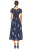 A woman viewed from the back wearing a navy midi dress with a white floral print, short puff sleeves, and a gathered waist, paired with navy heels against a plain white background.
